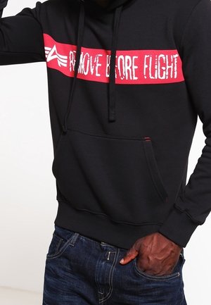 Czarna bluza z kapturem z czerwonym paskiem na wysokości klatki piersiowej, na którym znajduje się biały strzałka oraz napis „REMOVE BEFORE FLIGHT”, noszona z ciemnoniebieskimi dżinsami.