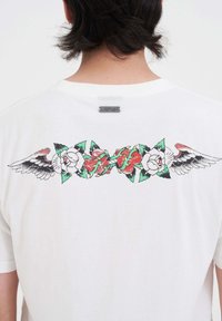 Espalda de un hombre que lleva una camiseta blanca con un diseño de alas y rosas al estilo de tatuaje en rojo, verde y negro en la parte superior de la espalda.