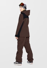 Brązowa kurtka narciarska z czarnymi akcentami, w połączeniu z pasującymi brązowymi spodniami. Model nosi szary beanie i gogle narciarskie. Luźny krój i funkcjonalny design.