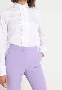 White blouse with ruffled front detail, paired with fitted lilac trousers. The blouse has a classic collar and button cuffs. Smooth fabrics.