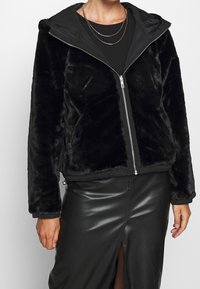 Svart faux fur-jacka med dragkedja fram, med en avtagbar huva, elastiska ärmar och en kort design. Kombination med en svart läderkjol.