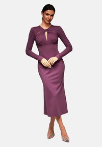 Kroppsnära lila klänning med lång ärm, nyckelhålsutskärning vid bröstet och elegant design, matchad med prydliga högklackade skor.