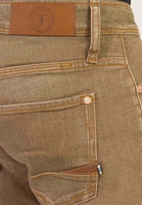 Bruine denim jeans met oranje stiksels, een leren label op de tailleband en meerdere zakken met gedetailleerde naden.