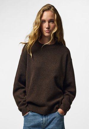 Jeune femme aux longs cheveux blonds portant un pull marron foncé et un jean bleu, debout les mains dans les poches devant un fond uni.