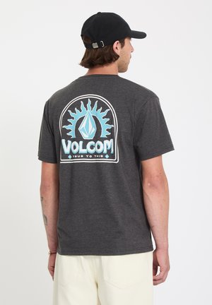 Mann trägt dunkles graues Volcom-T-Shirt mit blauem Sonnenlogo und Schriftzug "VOLCOM TRUE TO THIS" auf der Rückseite, schwarze Mütze und helle Hose, steht mit dem Rücken zur Kamera.