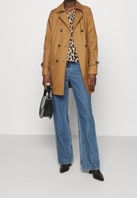 Osoba v hnědém dvouvrstvém kabátu, leopardím topu, modrých širokých džínách, černých botách a držící černou kabelku.