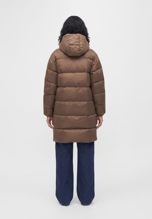 Personne aux cheveux bouclés foncés vue de dos, portant un long manteau matelassé marron avec une capuche et un pantalon bleu large.