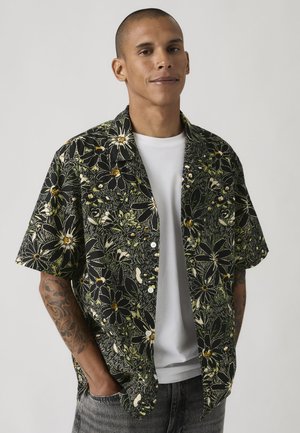 Hombre con una camisa negra de manga corta con flores y botones sobre una camiseta blanca, con las manos en los bolsillos, frente a un fondo liso.