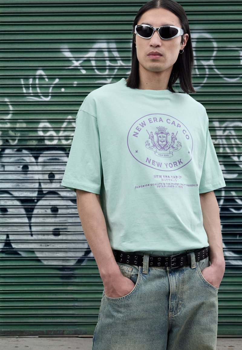 Light mint green t-shirt with purple graphic print, paired with light blue jeans and a black studded belt. Model wears white sunglasses.