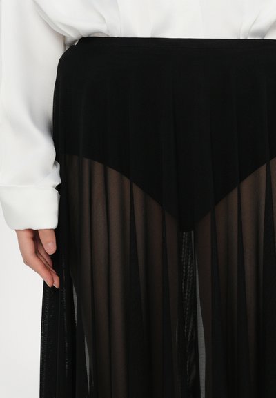 Falda negra transparente con capas de pliegues y cintura alta. Hecha de un material ligero y semitransparente. Acompañada de una blusa blanca de manga larga.