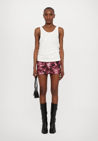 Witte tanktop met gedrapeerd detail, roze gemusterde minirok, zwarte kniehoge laarzen en een kleine zwarte handtas. Model staat tegen een effen achtergrond.