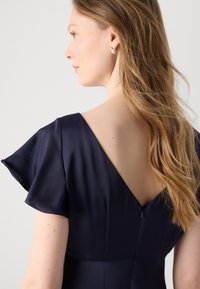 Rochie bleumarin învelitoare cu mâneci fluture, decolteu adânc la spate și închidere cu fermoar. Țesătură netedă cu un luciu subtil și o croială bine definită.