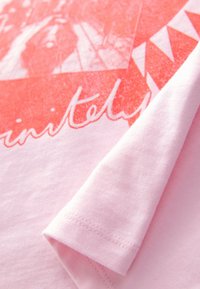 Svetlo roza bombažna majica s koralnim grafičnim vzorcem s trikotnimi vzorci in besedilom. Tkanina se zdi mehka s gladko teksturo.