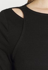 Musta ribattu neulepaita, jossa on pyöreä pääntie ja leikkaukset olkapäillä. Tekstuuri on pehmeä ja siinä on näkyviä ommelkoristeita.