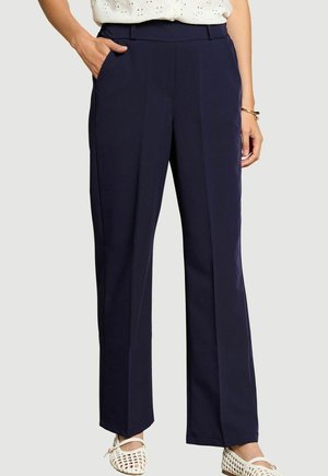 Mujer que lleva pantalones azul marino de pierna recta con bolsillos laterales, una blusa blanca con estampado, pulsera dorada y zapatos planos blancos tejidos.