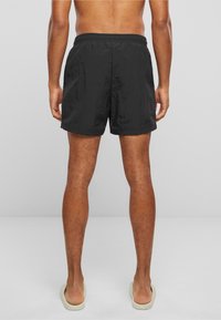 Shorts de bain noirs avec une taille élastique, fabriqués dans un tissu léger et texturé. Le mannequin porte des sandales beiges.