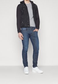 Chaqueta de mezclilla negra con capucha, camiseta gris, jeans azules y zapatillas blancas. La chaqueta tiene botones al frente y detalles desgastados.