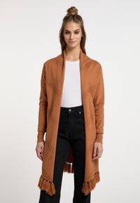 Lange cardigan in roestkleur, getextureerd met diagonale strepen en afgewerkt met een franjeszoom. Gemaakt over een aansluitend wit topje en zwarte broek.