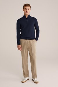 Maglione navy con zip, realizzato in materiale a maglia, abbinato a pantaloni beige a plissé e scarpe da ginnastica bianche con accenti marroni.