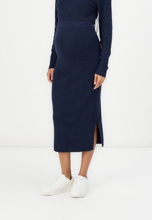 Granatowa dzianinowa sukienka midi z długimi rękawami, bocznym rozcięciem i dopasowanym fasonem. Noszona z białymi trampkami na jednolitym tle.