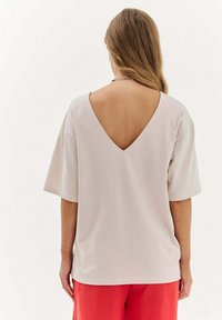 Světle béžové tričko s krátkými rukávy, designem ve tvaru V na zádech a jemnou strukturou. V párování s červenými kalhotami, které mají volný střih.