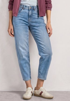 Person iført lyseblå korte jeans, cremefarvede sneakers med brune striber og en mørkerød lynlåsjakke over en hvid skjorte.