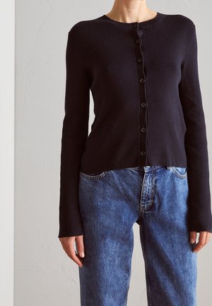 Osoba ubrana w czarny prążkowany kardigan z guzikami i wysokim stanem niebieskich dżinsów, stojąca na tle gładkiej jasnej ściany.