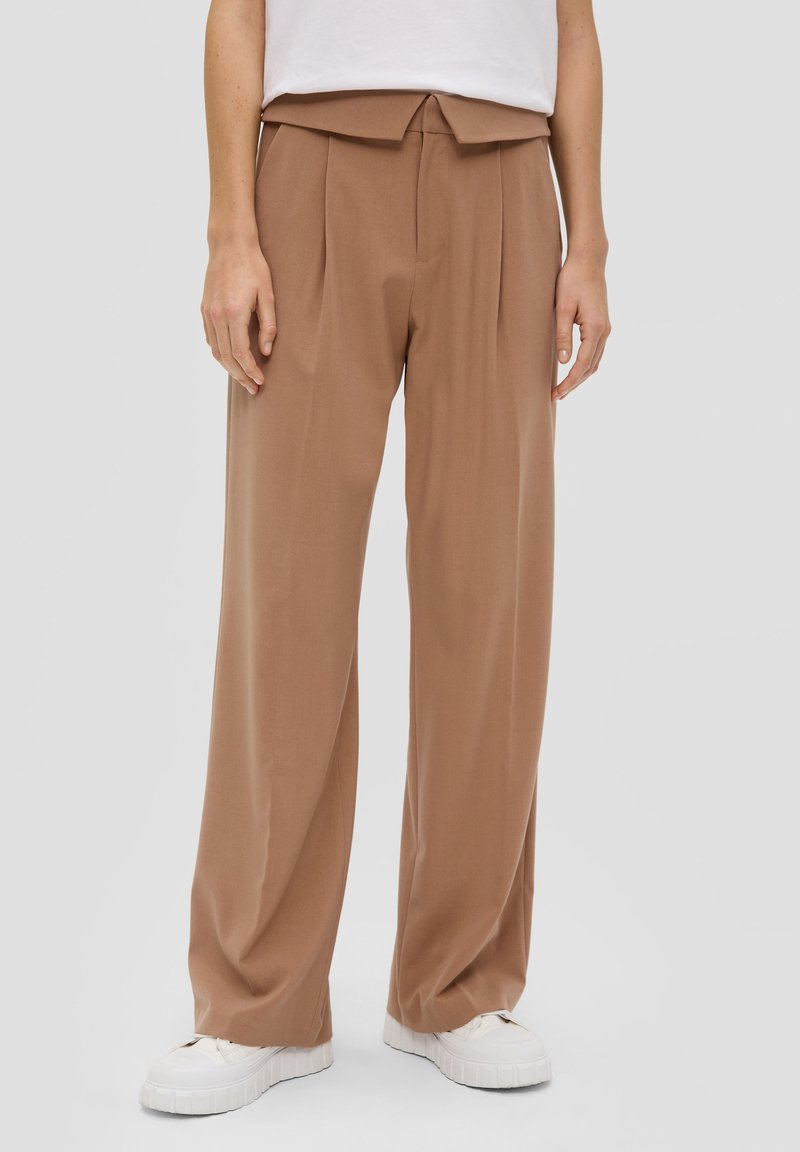 Pantalons marron à jambe large avec des plis et une taille nouée. Tissu lisse avec une finition mate, associés à des baskets plateformes blanches.