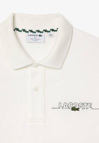 Polo bianca con texture a coste, dotata di due bottoni, logo ricamato "LACOSTE" e piccolo emblema di coccodrillo verde sulla parte sinistra.