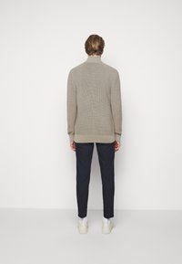 Beige stickad tröja med hög krage, ribbad textur och långa ärmar, ihop med mörka byxor och ljusa skor.