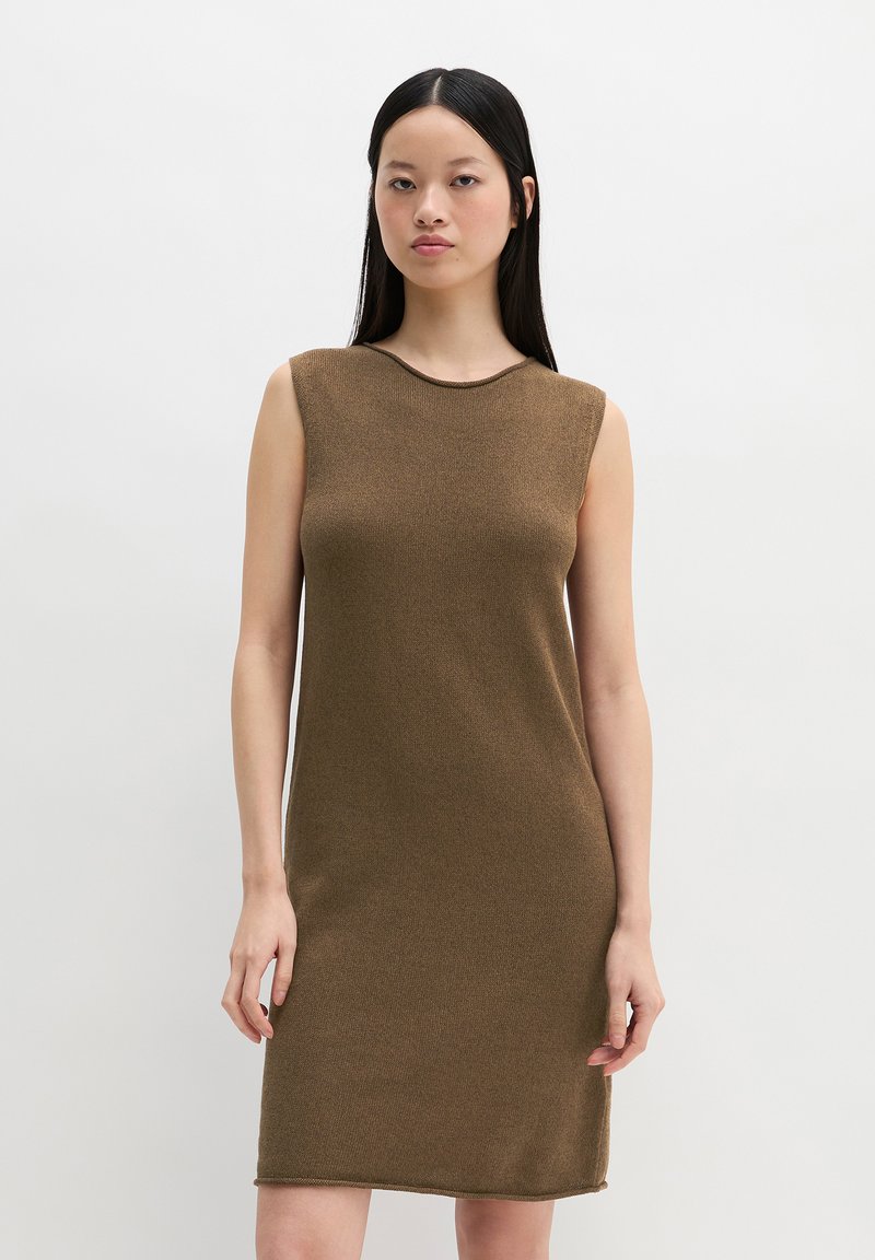 Frau mit langen schwarzen Haaren, die ein ärmelloses, knielanges braunes Strickkleid trägt und vor einem schlichten weißen Hintergrund steht.