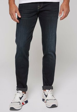 Dark blue denim jeans, fitted design, five pockets, subtle stitching details, cuffed hems, paired with white and navy athletic sneakers.