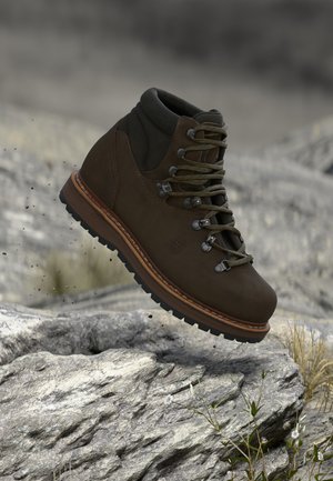 Brown hiking boot with rugged sole hovering above rocky terrain with sparse grass and small white flowers.