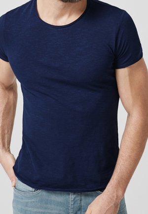 Homme portant un t-shirt bleu foncé ajusté à manches courtes et un jean bleu clair, debout avec une main dans la poche devant un fond uni.