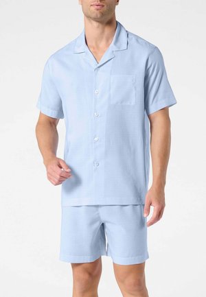 Uomo che indossa una camicia azzurra a maniche corte con taschino sul petto e pantaloncini abbinati su uno sfondo semplice.