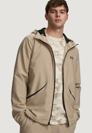 Giovane uomo che indossa una giacca beige con cappuccio e zip sopra una camicia mimetica chiara, in piedi con le mani nelle tasche su sfondo neutro.