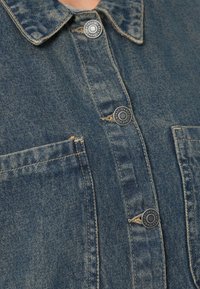 Jeans srajca z ovratnikom, temno modre barve, dvema žepoma na prsih in kovinskimi gumbi z embosiranimi detajli. Teksturirana površina.