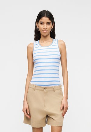 Nő világoskék-fehér csíkos ujjatlan felsőben és bézs rövidnadrágban, egyszínű fehér háttér előtt áll.