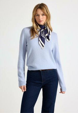Vrouw draagt lichtblauwe trui, donkerblauwe jeans en een marineblauw-wit gestreepte sjaal om haar hals gebonden, staand tegen een effen achtergrond.