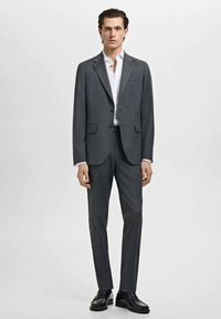 Traje gris a medida con chaqueta de un solo botón, solapas en muesca y pantalones planos. Combinado con una camisa blanca y zapatos negros.