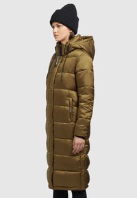 Manteau matelassé long couleur olive avec capuche, design matelassé, fermeture éclair à l'avant, cordons réglables et poches latérales, porté avec un bonnet en tricot noir.