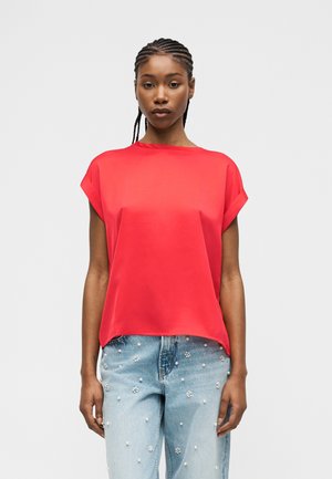 Femeie cu păr împletit, purtând o bluză scurtă cu mâneci roșii aprinse și blugi bleu deschis decorați cu mărgele de perle și flori.