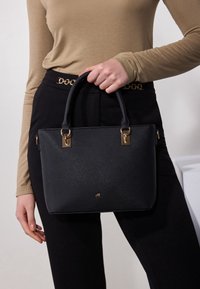 Personne tenant un sac à main noir texturé avec des détails dorés, portant un haut à manches longues beige et un pantalon noir avec une ceinture chaîne dorée.