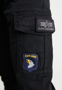 Crne cargo hlače s oznakom "Alpha Industries" i amblemom orla označenim "AIRBORNE", s džepovima sa strane.