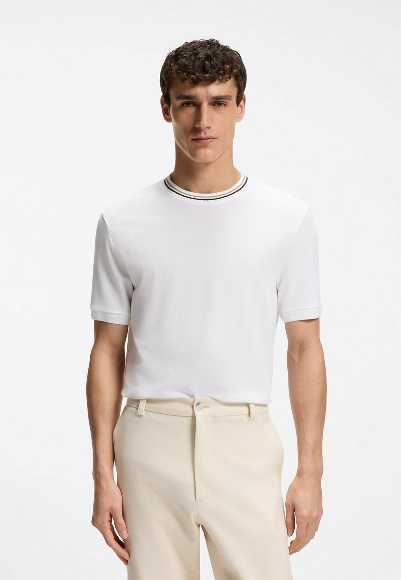 Homme portant un pull à manches courtes blanches avec un col rond rayé, rentré dans un pantalon beige, debout devant un fond uni.