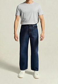 Jean en denim bleu foncé à coupe ample, avec un design classique à cinq poches et une ceinture standard. Associé à un t-shirt gris clair.