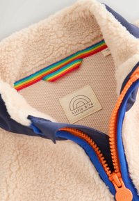 Jasnobrązowa, pluszowa kurtka polarowa z granatowym kołnierzem i pomarańczowym zamkiem błyskawicznym. Posiada wielokolorowy paskowany detal oraz metkę wewnątrz.