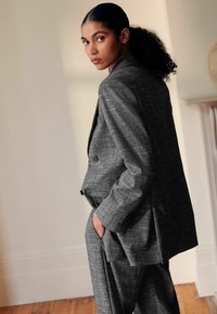 Grå tekstureret jakkesæt med en dobbeltbrystet blazer, der har store knapper og skræddersyede bukser. Modellen står med hånden i lommen i en neutral setting.