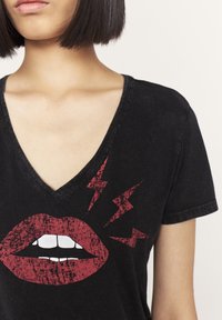 Musta V-kaula-aukkoinen T-paita, jossa kuluneet punaiset huulet valkoisilla hampailla ja vasemmalla punaiset salamaniskut, lyhyet hihat ja rento istuvuus.