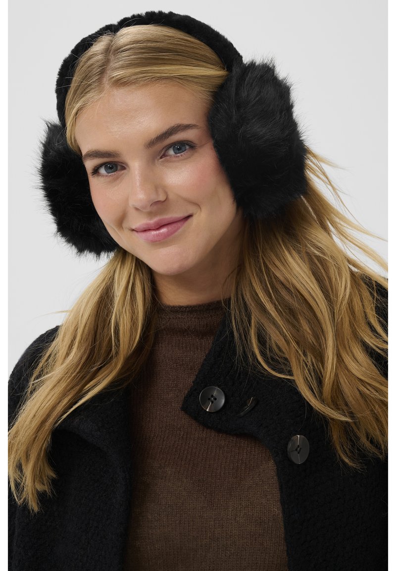 Femme aux longs cheveux blonds portant des cache-oreilles noirs en fourrure, un manteau sombre à boutons et un pull marron, souriant légèrement.
